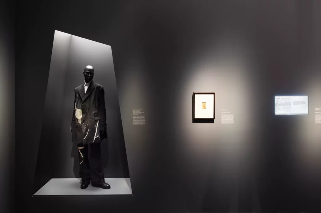 A gallery display showing a mannequin in a black ensemble and a framed picture on a wall