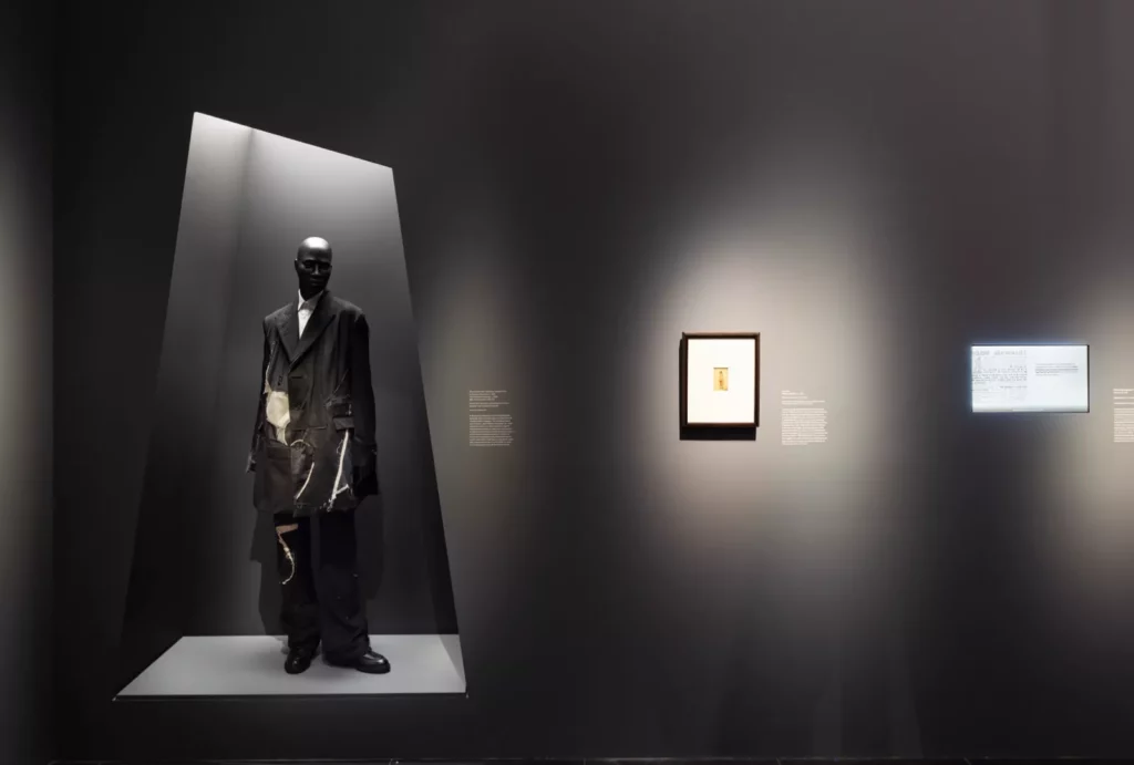 A gallery display showing a mannequin in a black ensemble and a framed picture on a wall