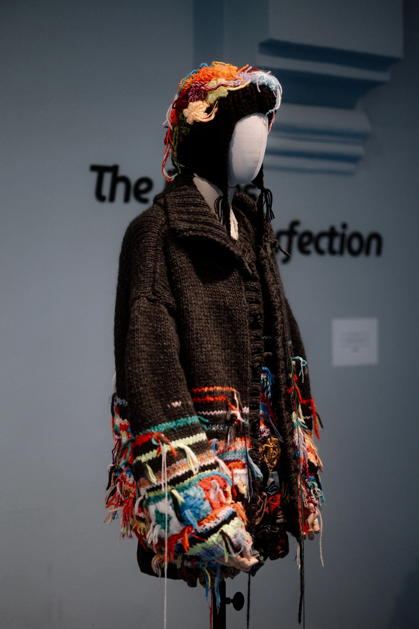 Mannequin wearing an oversized dark knitted cardigan with colourful crochet patchwork details and a multicoloured yarn headpiece