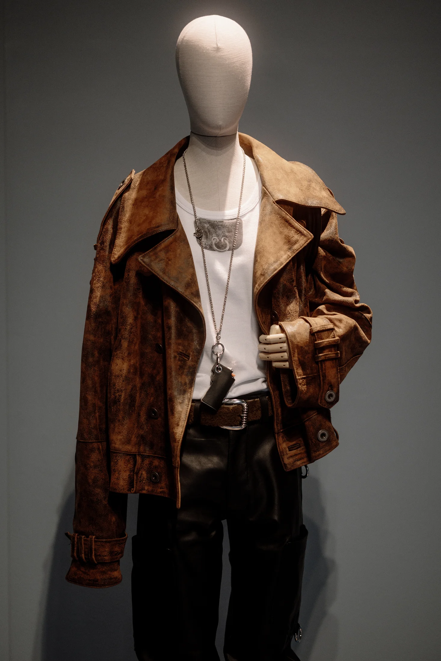 Mannequin in a distressed brown leather biker jacket, white t-shirt, chain necklace with small pouch, and black leather trousers