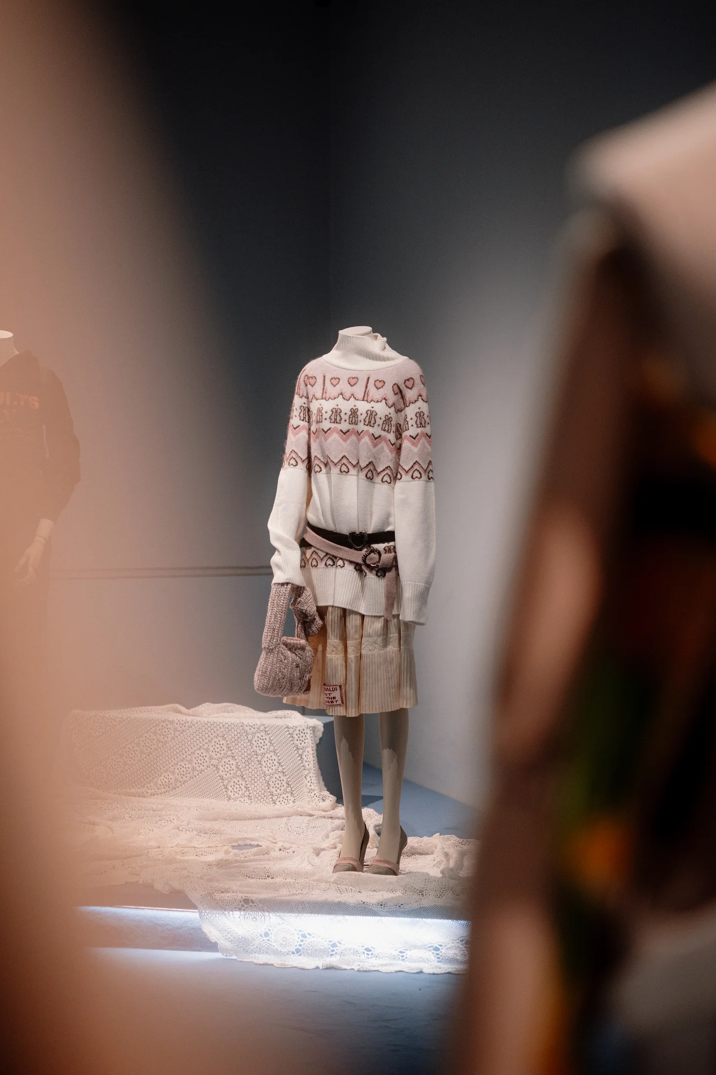 Mannequin in a cream fair isle knit jumper belted over a pleated skirt, holding a small knitted bag