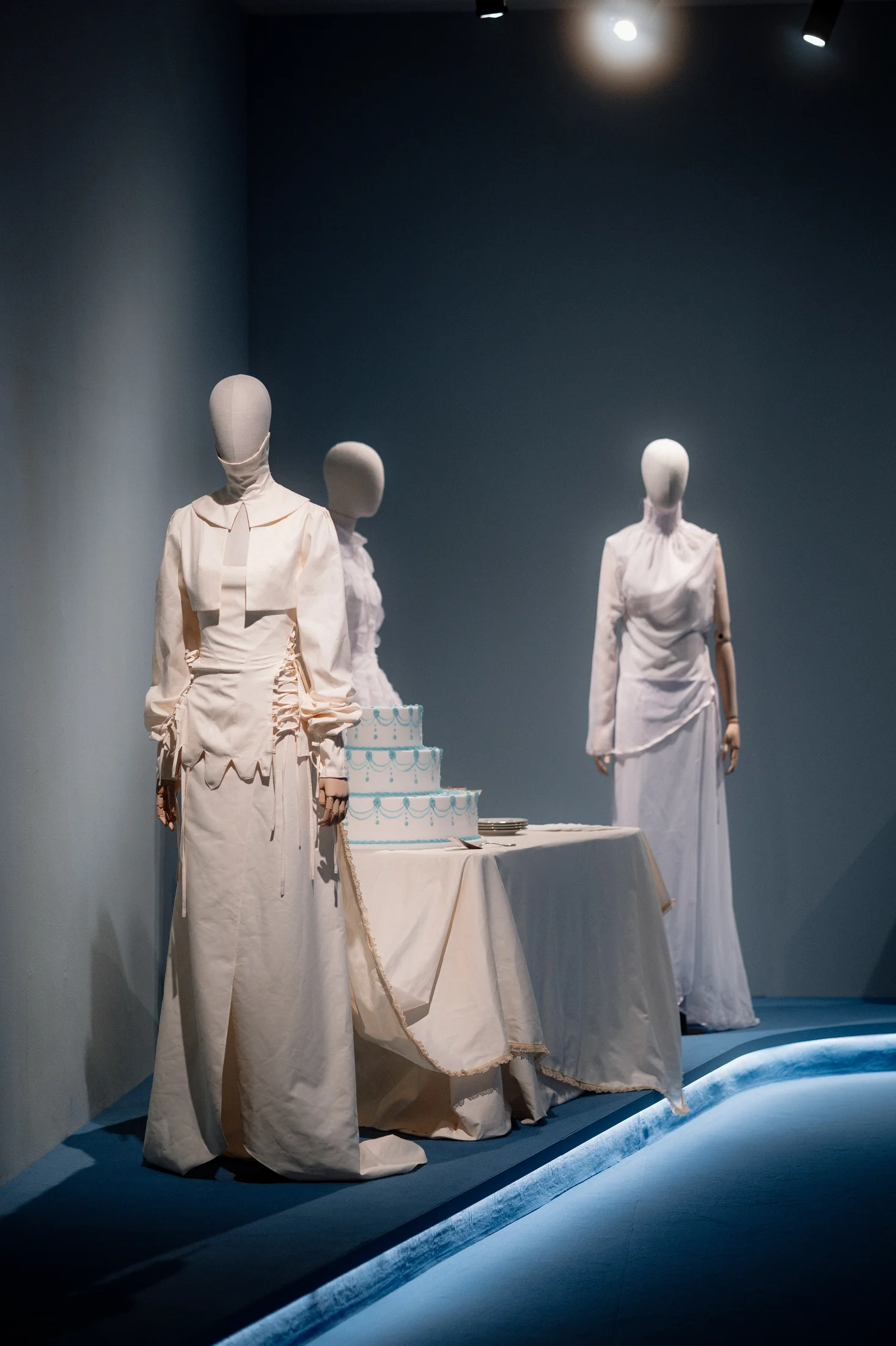 Three mannequins in white bridal-inspired garments alongside a tiered blue and white wedding cake on a draped table