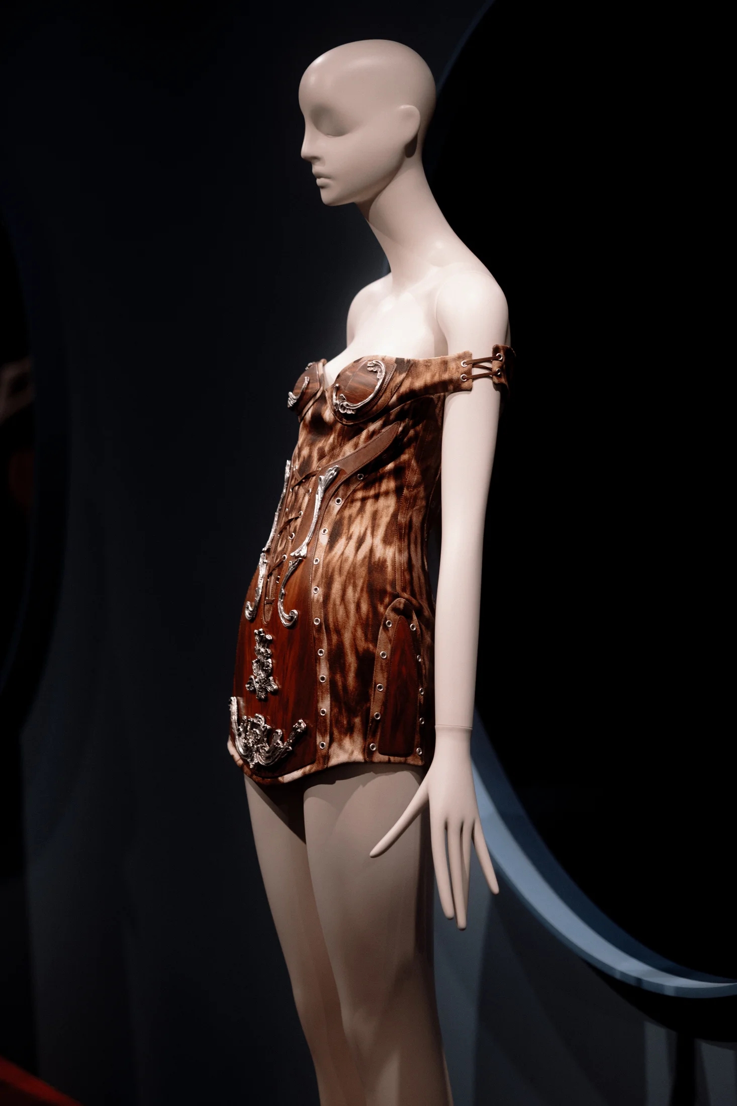 Close-up of a mannequin in a brown off-shoulder mini dress with silver metallic ornamental details and grommets