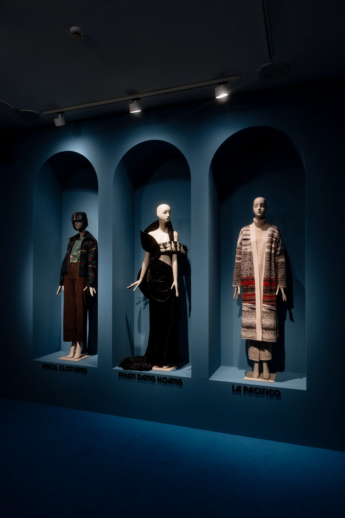 Three mannequins displayed in arched alcoves against a blue wall, wearing designs by Pikol Clothing, Phan Dang Hoang, and La Pacifico