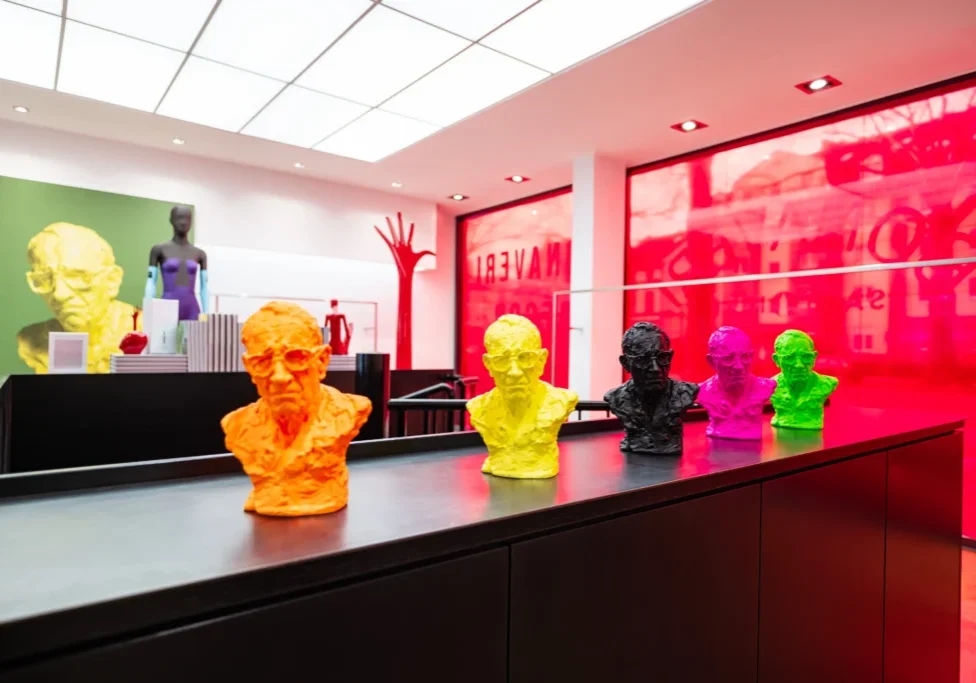 A row of brightly coloured miniature busts from Bonaveri's The 10 Collection displayed on a dark counter, with the signature red vinyl-covered showroom windows visible behind, at EuroShop 2026 in Dusseldorf.