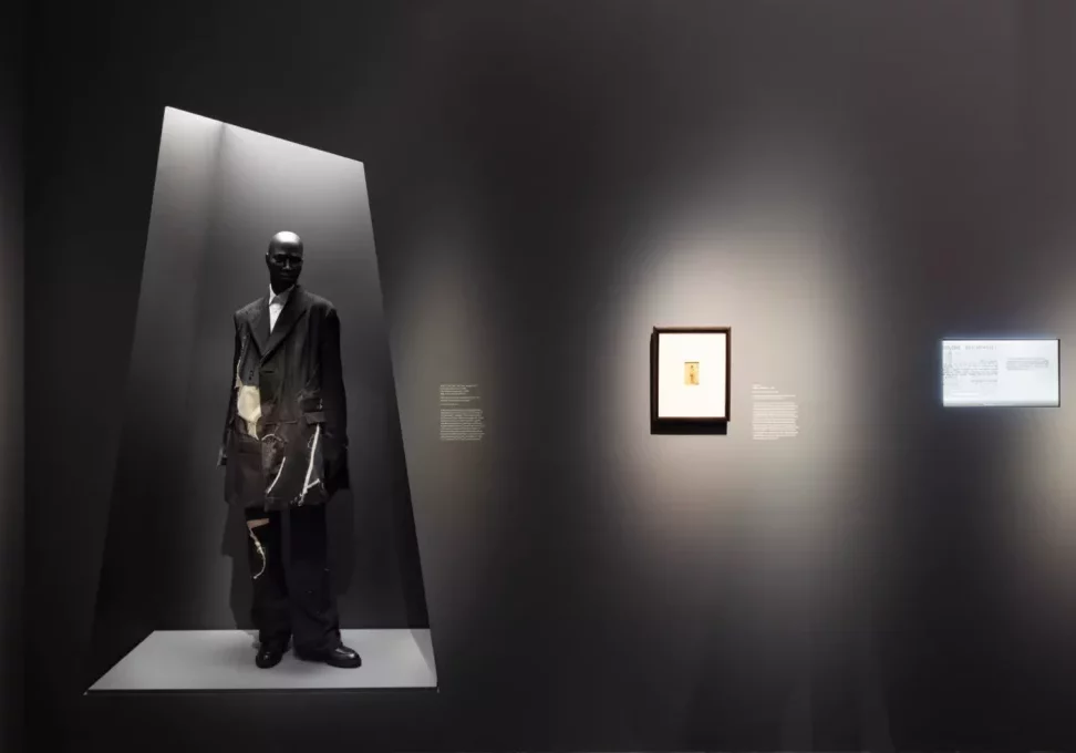 A gallery display showing a mannequin in a black ensemble and a framed picture on a wall