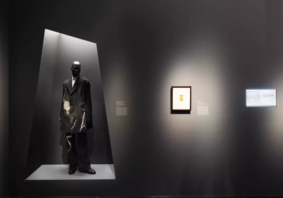 A gallery display showing a mannequin in a black ensemble and a framed picture on a wall