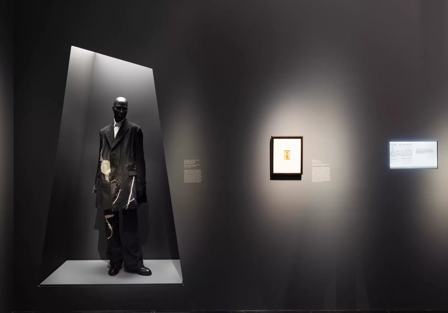 A gallery display showing a mannequin in a black ensemble and a framed picture on a wall