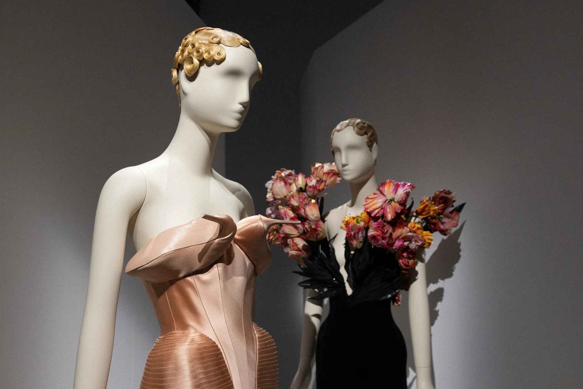 Two mannequins with sculpted gold hair, one in a pleated peach gown holding a bouquet of dried roses