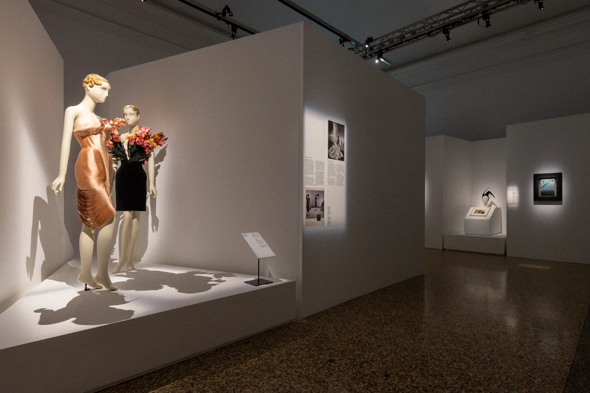 Gallery corner with two mannequins in a peach gown and black dress, framed photographs on the walls