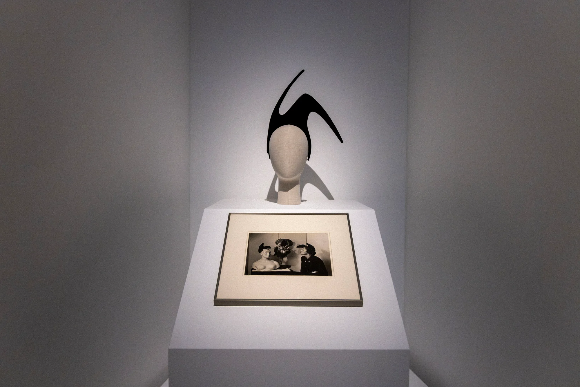 Mannequin head wearing a black shoe-shaped hat above a framed vintage photograph on a white plinth