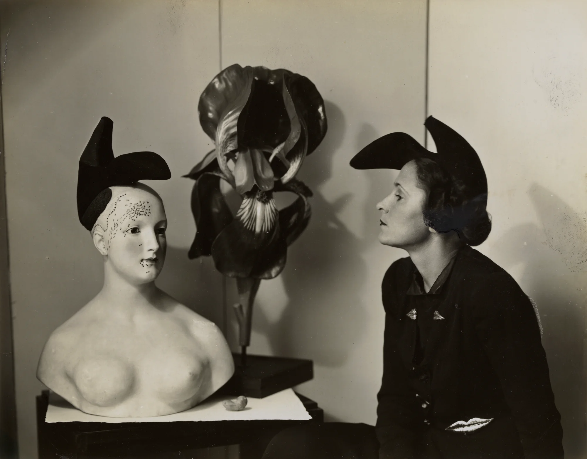 Black-and-white photograph of a woman beside a plaster bust wearing a shoe hat, with a large flower