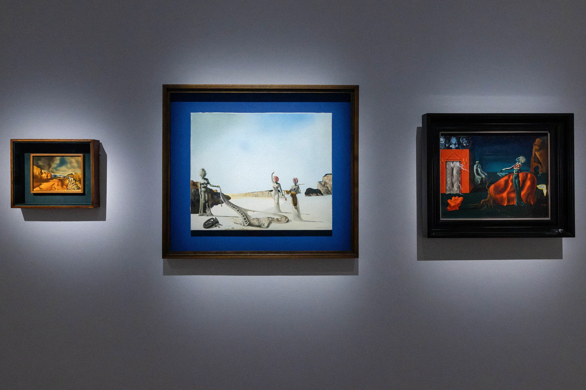 Three surrealist paintings hung on a grey gallery wall under spotlights