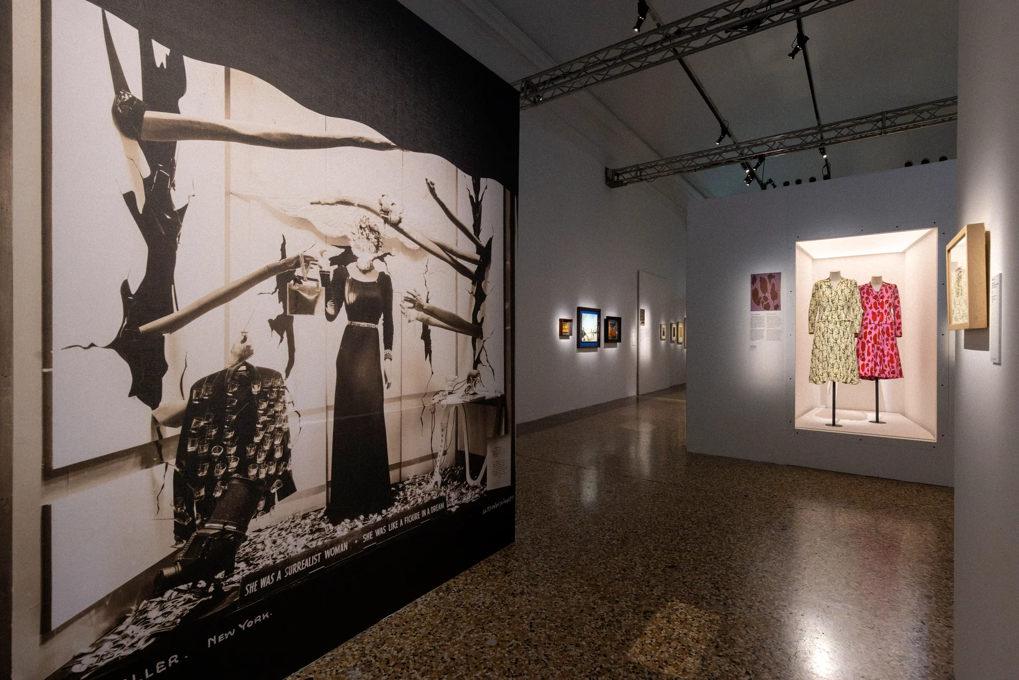 Large black-and-white mural of figures in a surreal landscape, two printed dresses in a lit niche beyond