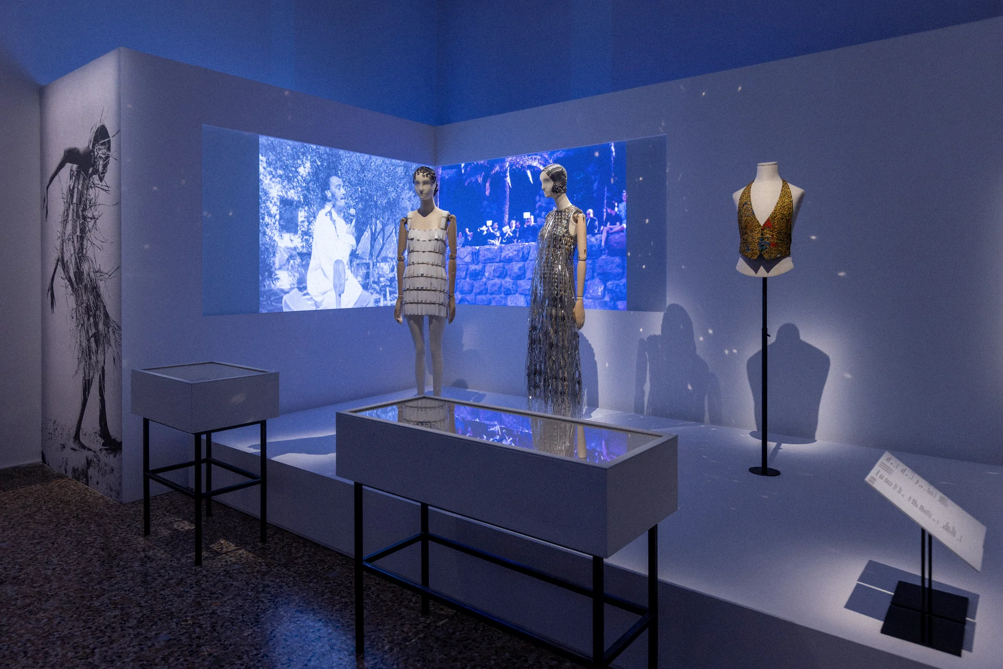 Blue-lit gallery room with a video projection, mannequins in sequinned dresses and a glass display case