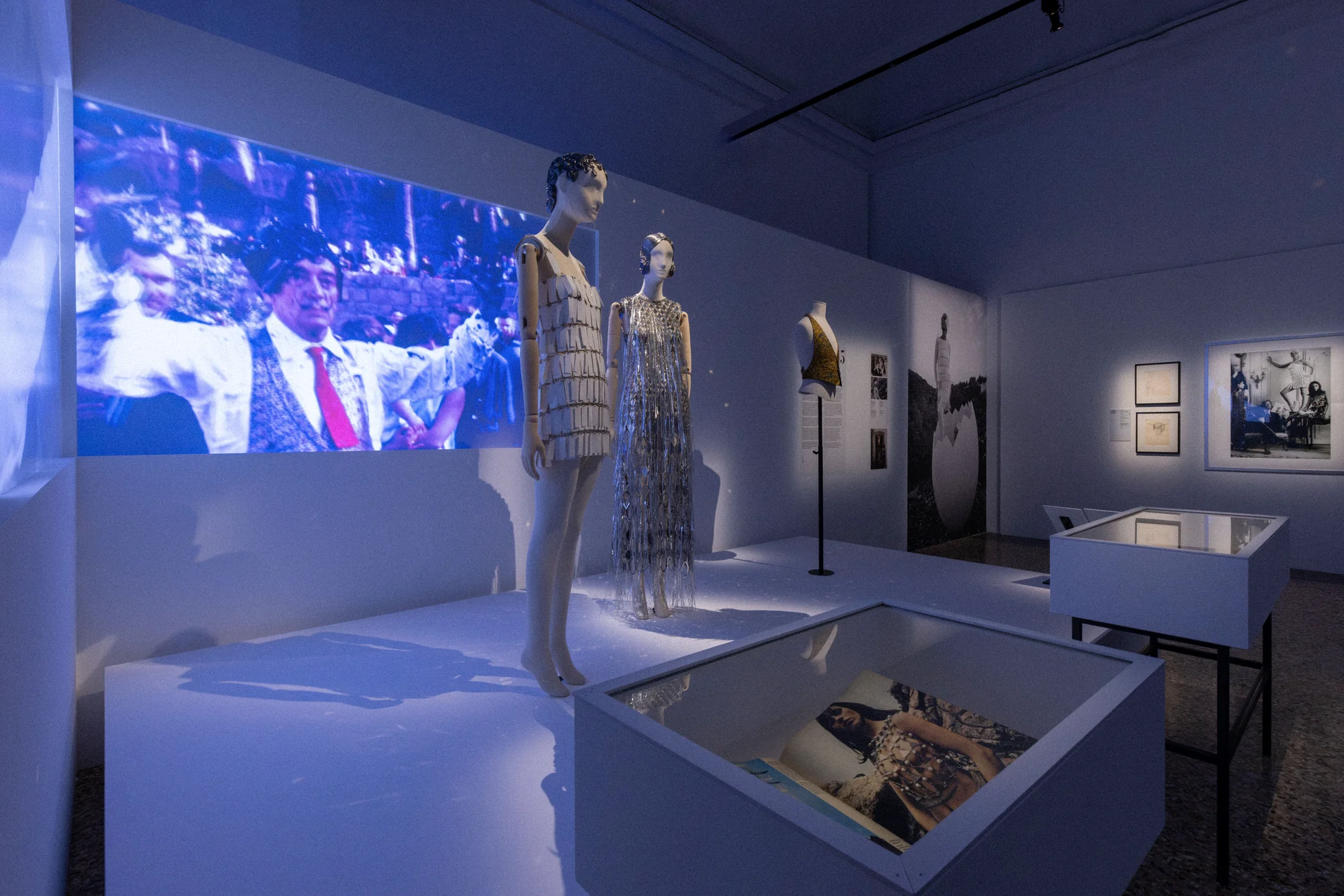 Mannequins in beaded dresses in front of a projected film, with display cases and framed works