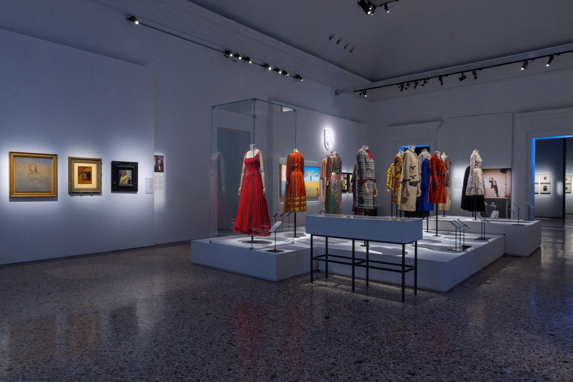 Wide view of the exhibition hall: a long plinth of dresses and coats, paintings on the walls