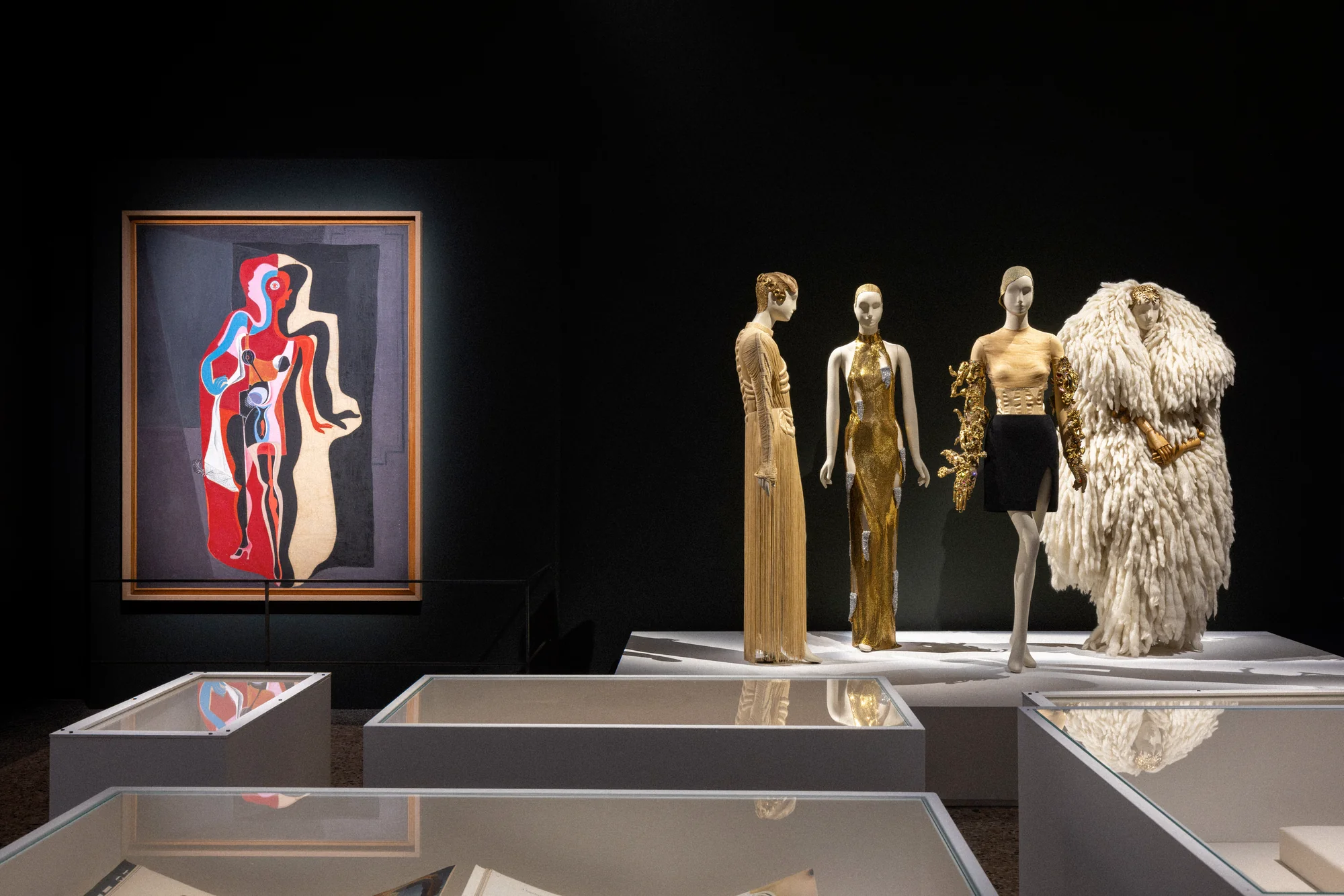 Dark room with a surrealist painting of a colourful figure and mannequins in gold outfits and a feathered coat