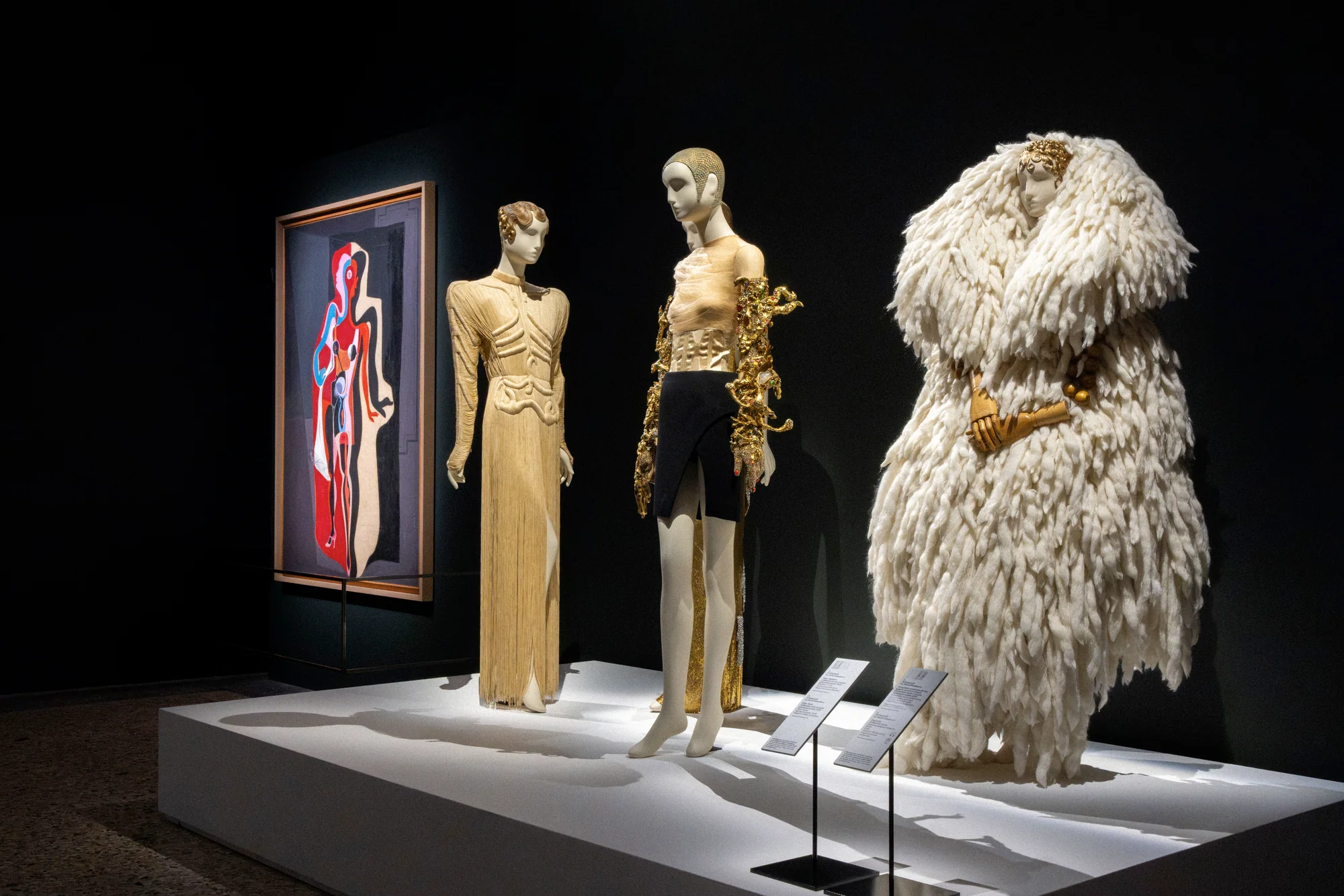 Mannequins in a gold gown, gold top with black skirt and a white feathered coat beside a surrealist painting