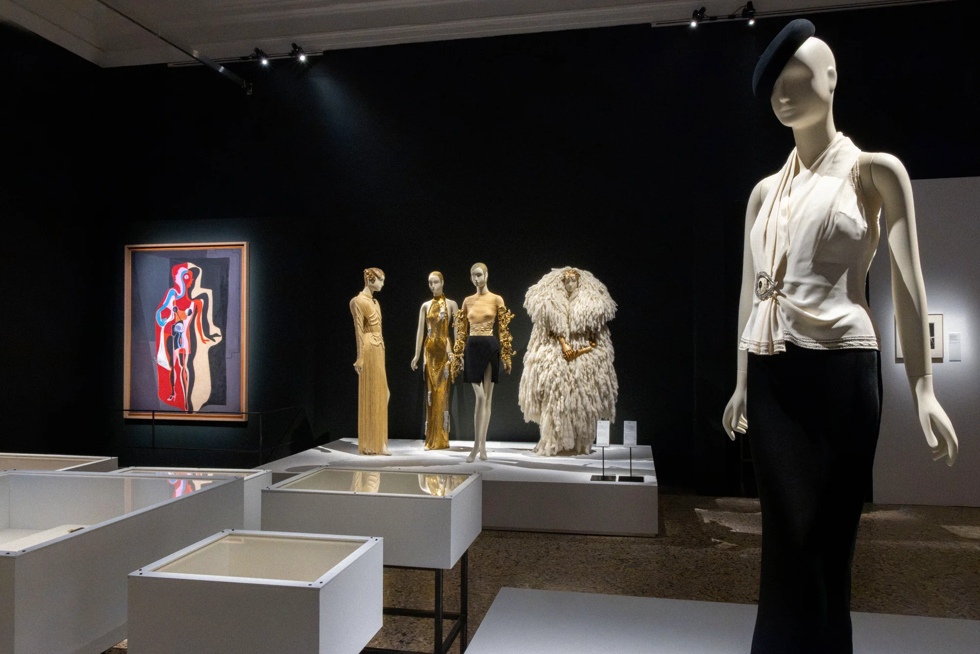 Mannequin in a white halter top, black trousers and black hat, gold outfits and a painting behind