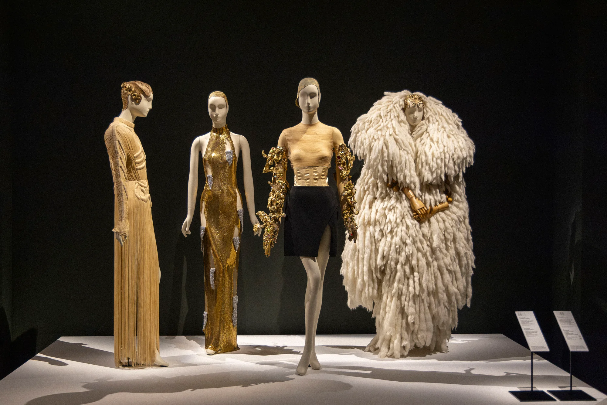 Four mannequins in gold gowns, a gold top with black skirt and a white feathered coat on a dark stage