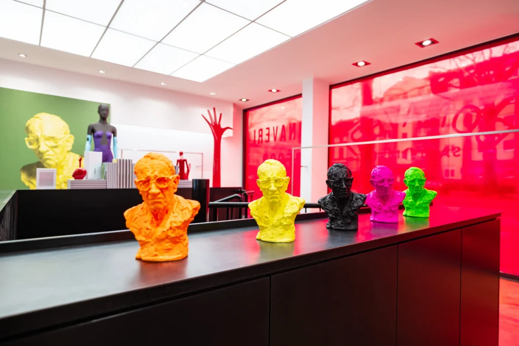 A row of brightly coloured miniature busts from Bonaveri's The 10 Collection displayed on a dark counter, with the signature red vinyl-covered showroom windows visible behind, at EuroShop 2026 in Dusseldorf.