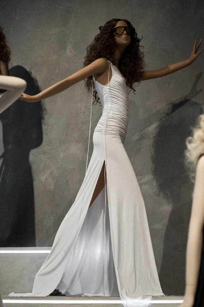 Mannequin with curly hair and sunglasses in a white draped gown against a grey wall