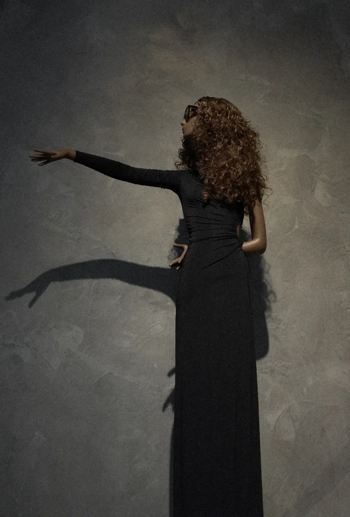 Mannequin with curly hair in a long black dress, one arm stretched along a grey wall