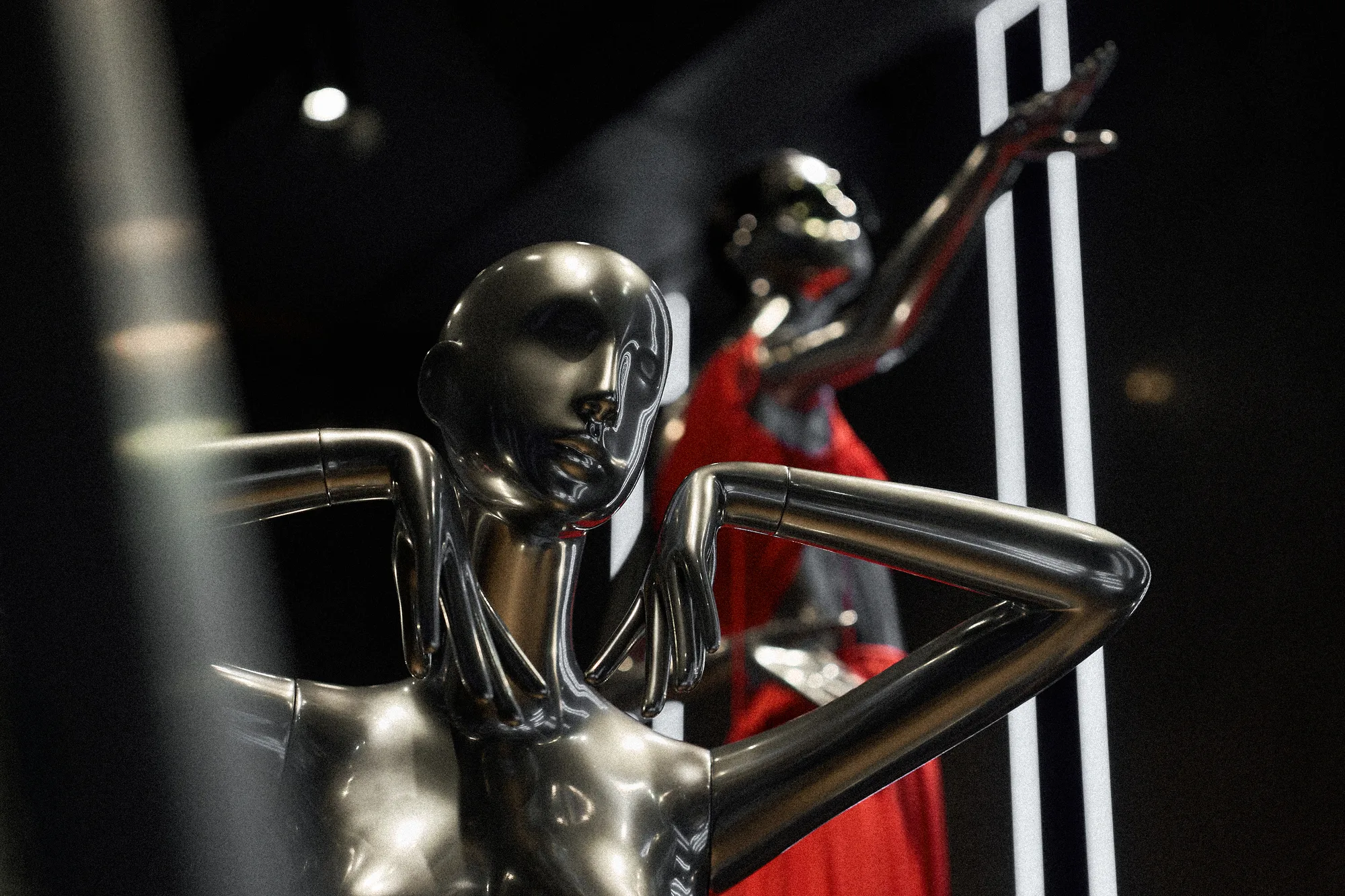 Chrome mannequin with one arm raised to its head, a red gown and light strips behind