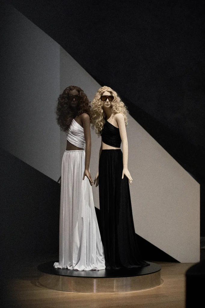 Two mannequins on a round plinth, one in white and one in black, with curly and blonde wigs