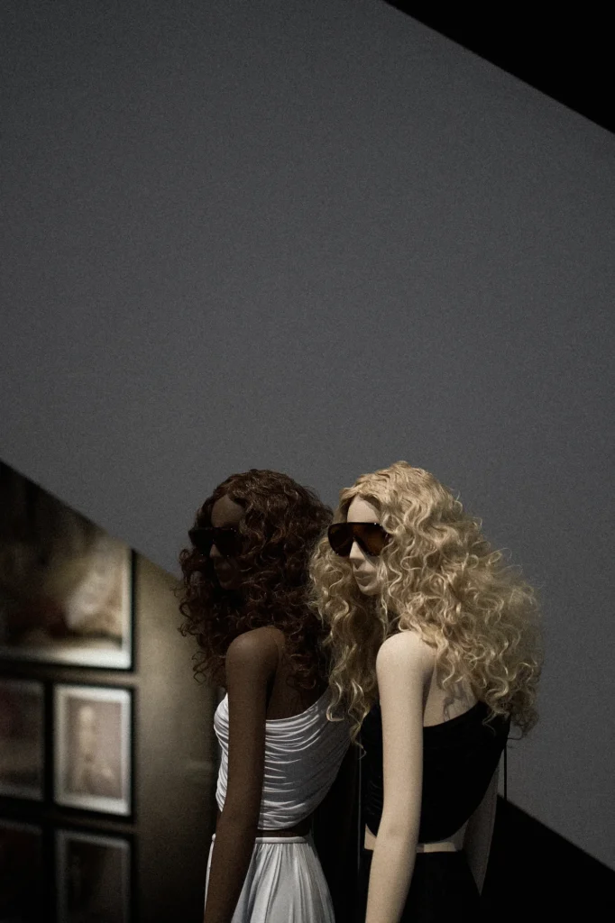 Two mannequins with curly wigs in white and black in front of archive photographs