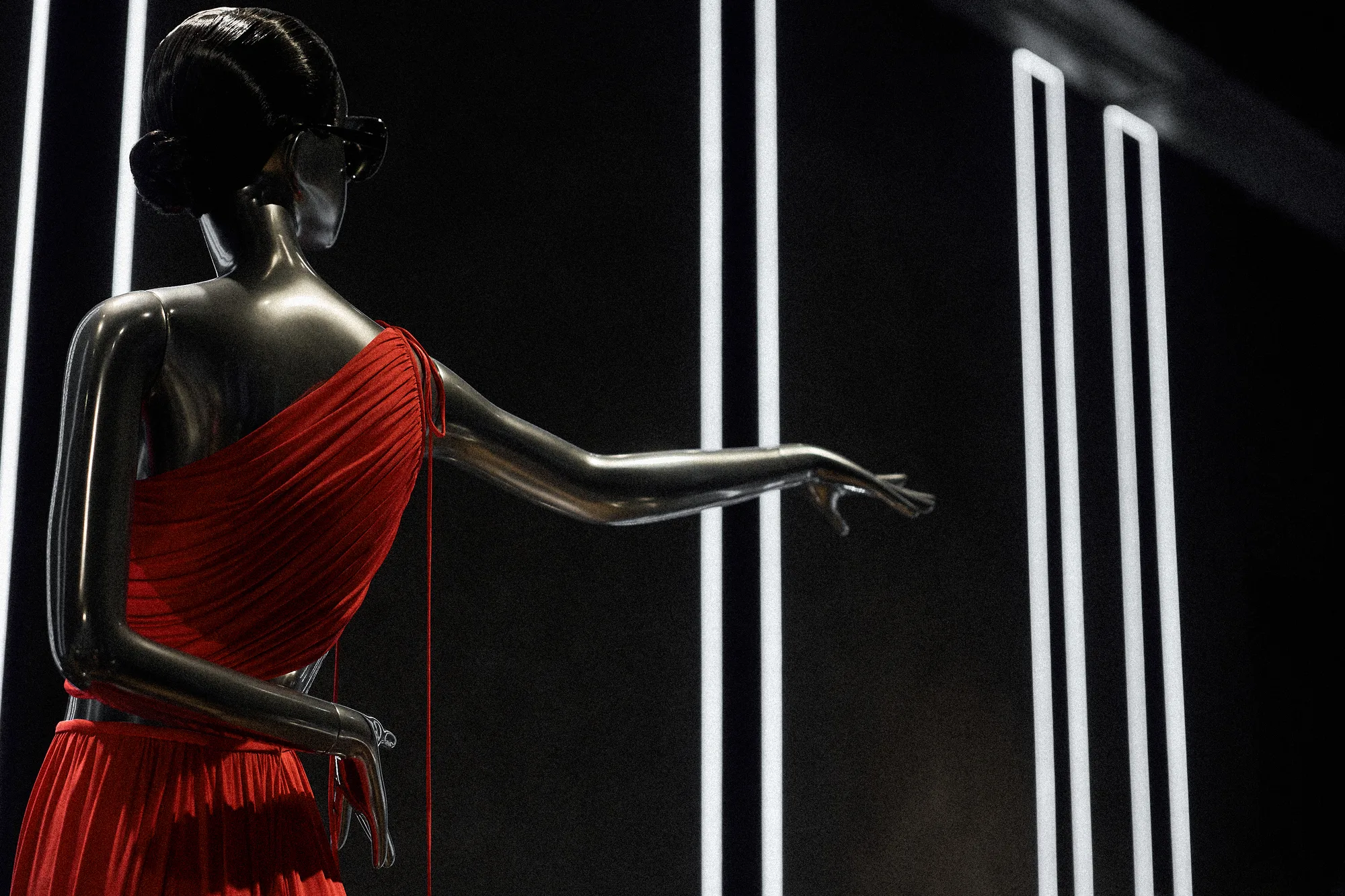 Chrome mannequin seen from behind in a red draped one-shoulder dress, white light strips beyond