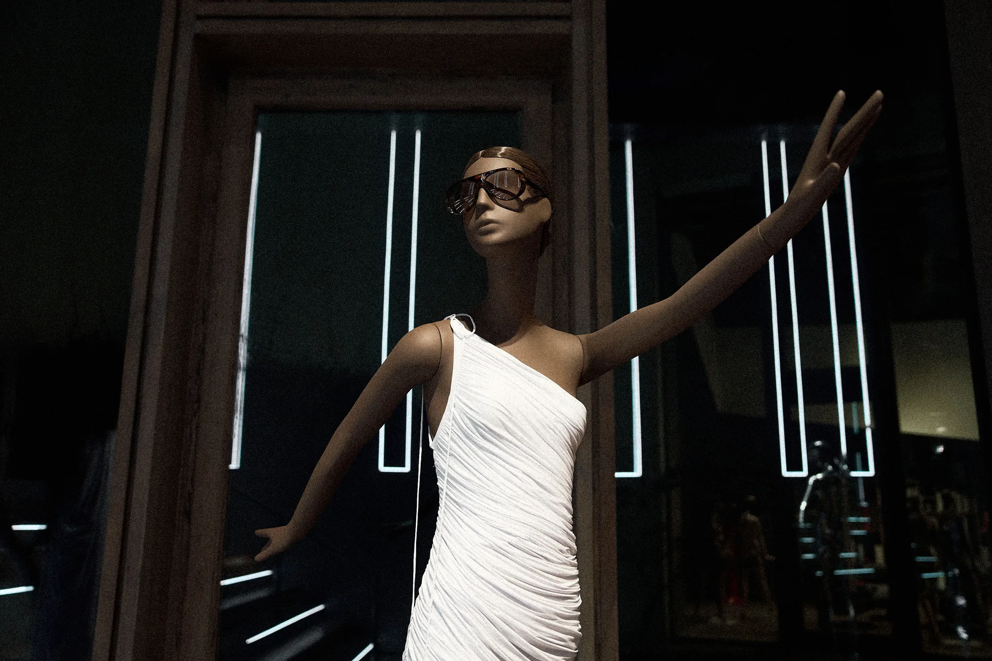 Mannequin in a white one-shoulder dress and sunglasses with one arm raised, framed by light strips