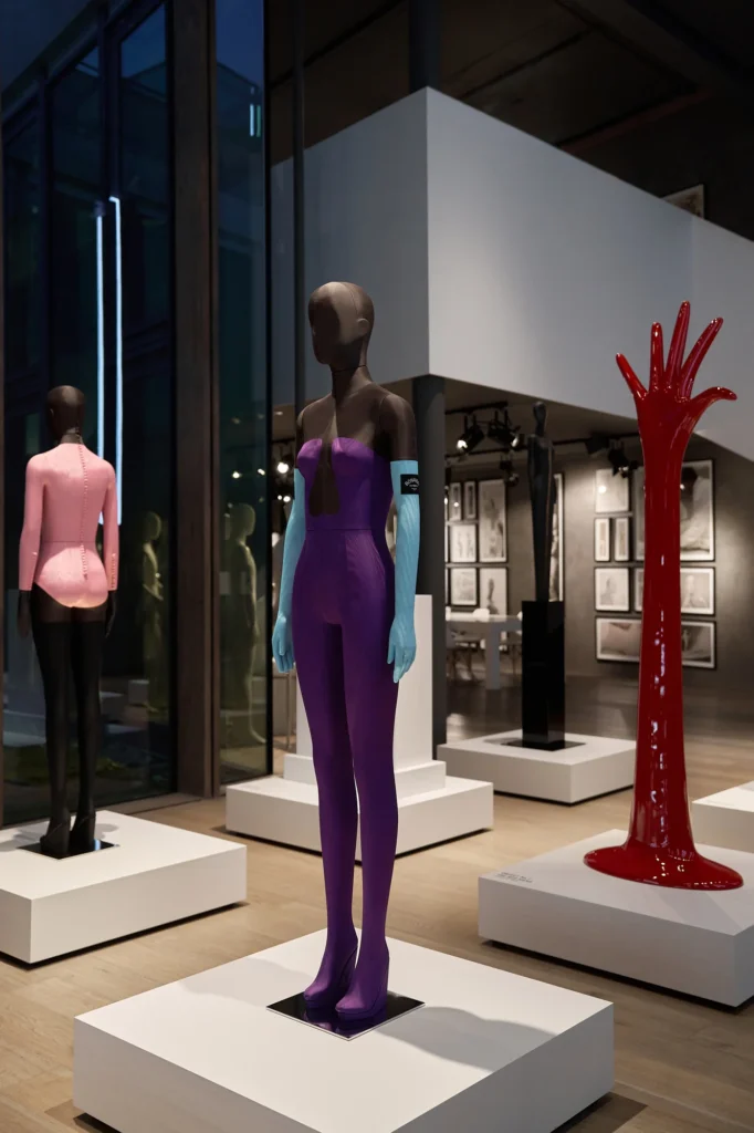 Mannequin in a purple strapless gown and blue gloves, with a pink-jacketed mannequin and a red hand sculpture behind